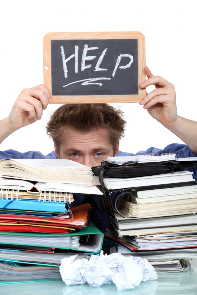 Student behid a stack of books, holding up a "Help" sign.